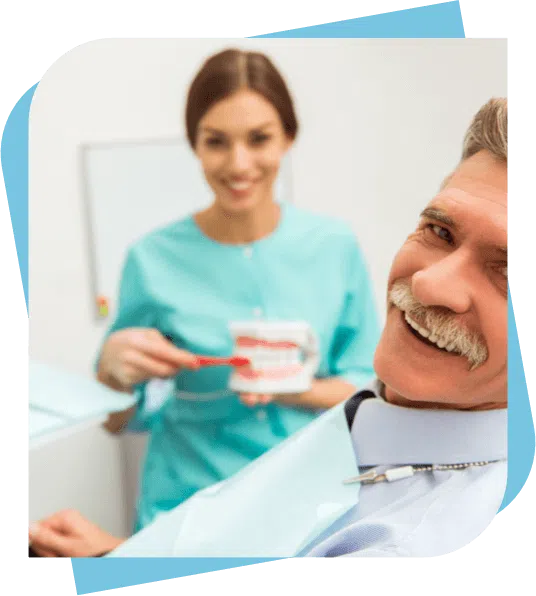 Middle aged man smiling while the dentist shows proper brushing techniques on a model in the backgorund.