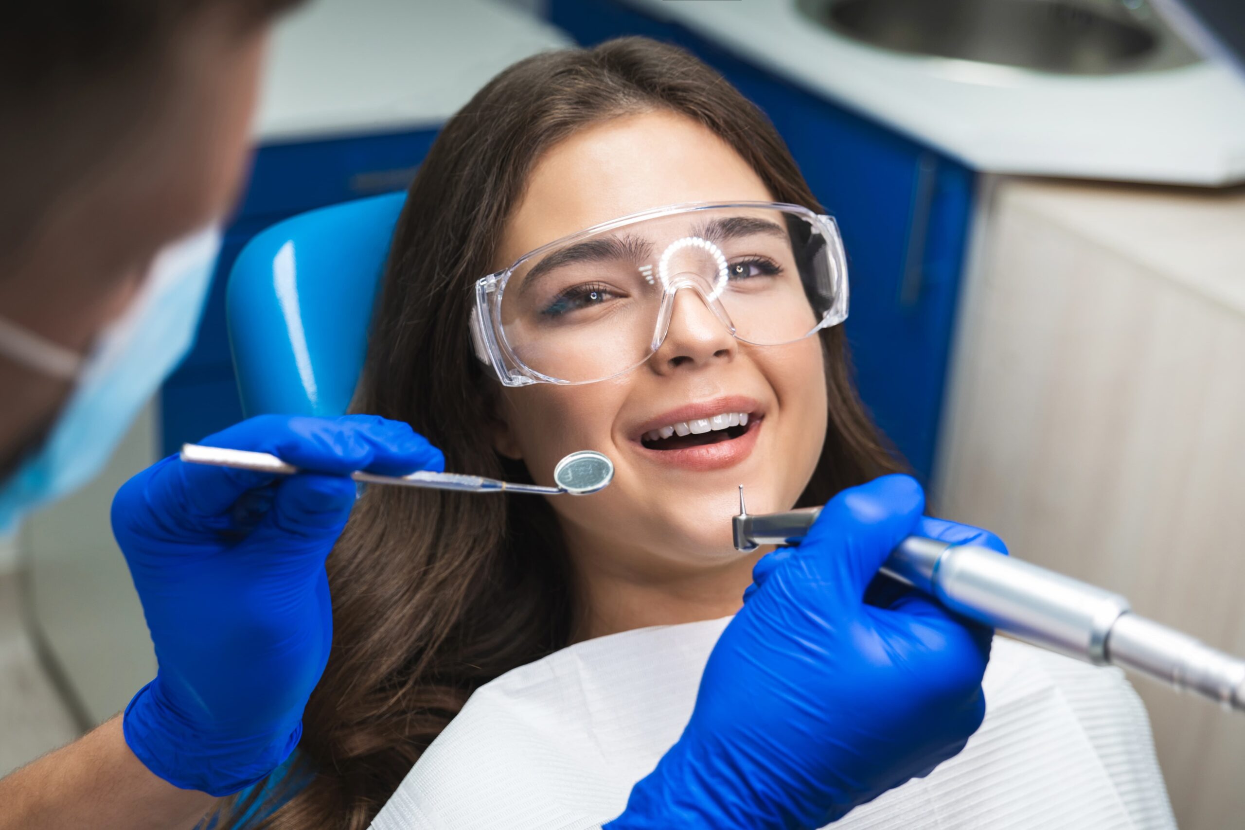 A young woman in a dentists chare with clear plastic glasses and a dental bib smiles up at the camera while a dentist in blue gloves reaches in with a mirror and scaler.