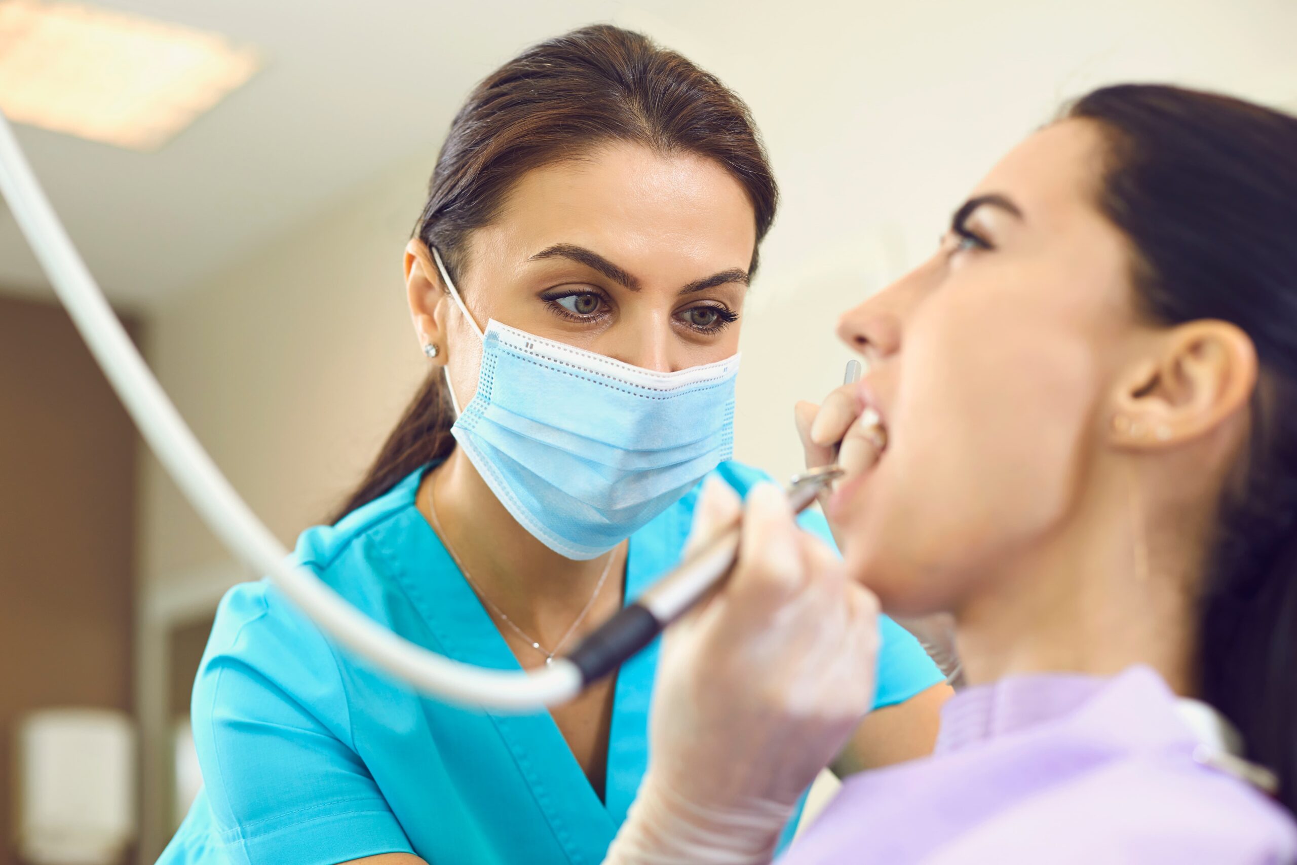 A dentist in scrubs and a blue facemask uses a tool connected to a tube on a woman sitting with her mouth open.