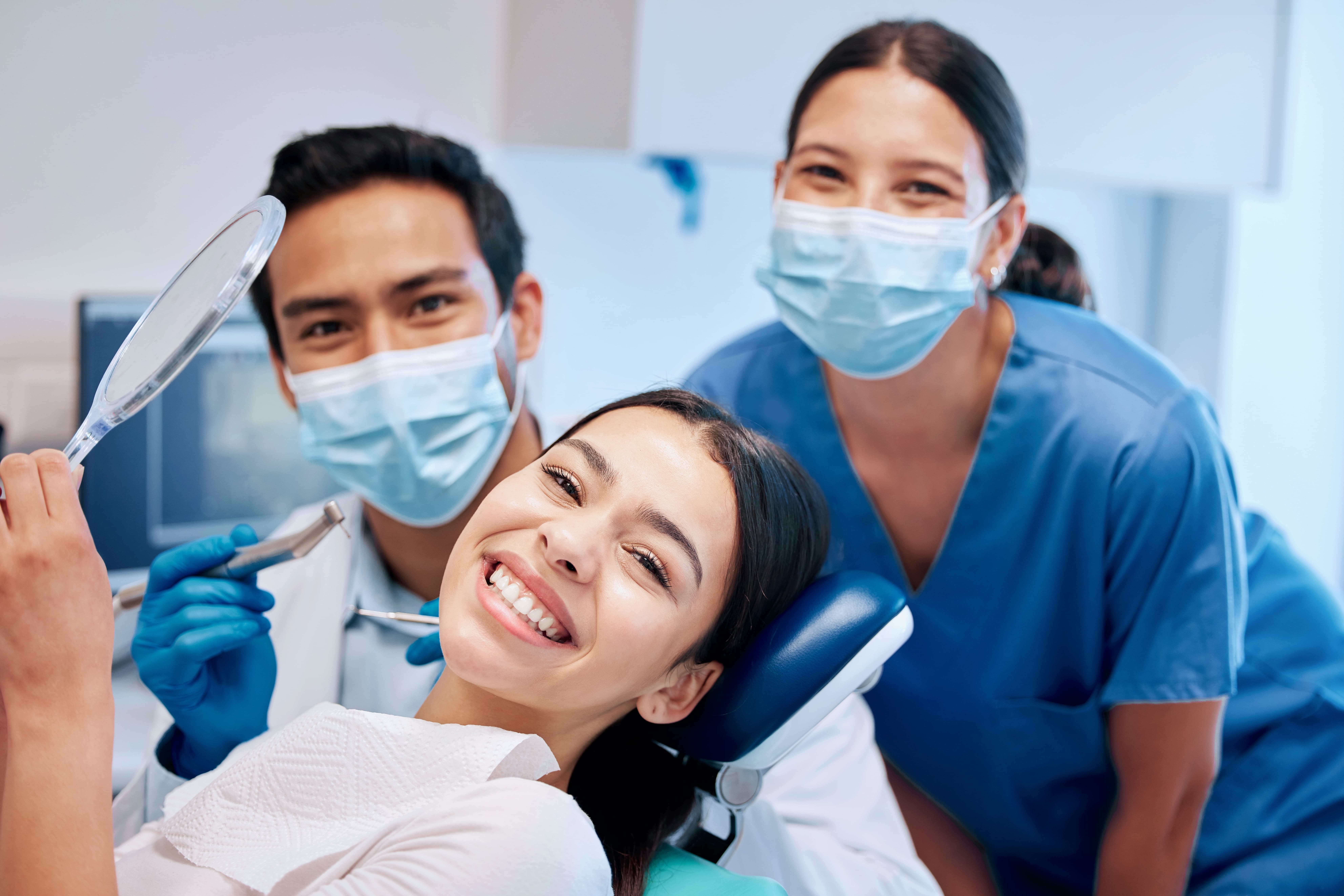 A girl on a dentist chair smiles at the camera. Behind her, a male dentist and female dental hygienist smile while wearing blue masks.