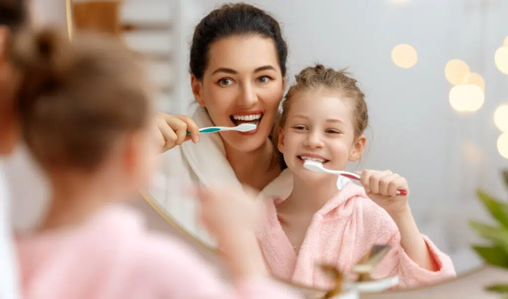 A mother and daughter brush their teeth together in a mirror.